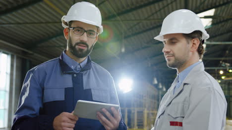 two workers wearing helmets talking while looking at the tablet in a big factory, then they look at the camera