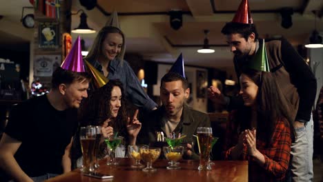 un joven guapo está haciendo deseos y soplando velas en el pastel mientras celebra su cumpleaños en el bar con amigos. la gente feliz lo está felicitando y aplaudiendo.