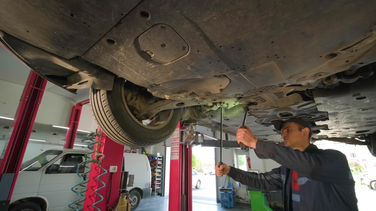 Young car mechanic at repair service station inspecting car wheel and suspension detail of lifted automobile. Bottom view.