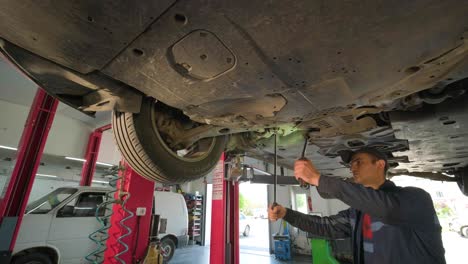 joven mecánico de automóviles en una estación de servicio de reparación inspeccionando la rueda del automóvil y el detalle de la suspensión de un automóvil levantado. vista de abajo.