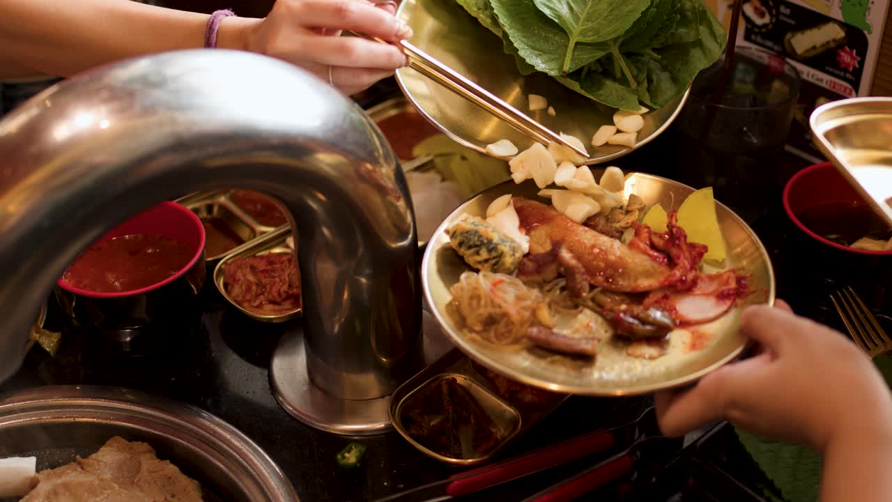 Diner uses tongs to add garlic to assorted Korean BBQ meats under warm, ambient lighting