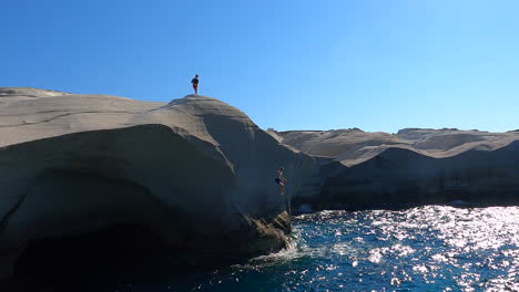 two people stand on the highest point of a cliff, one jumps into the water of the mediterranean sea