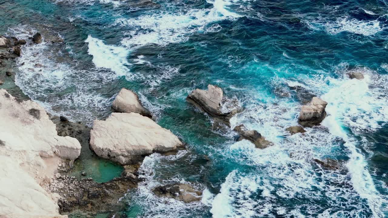 Gentle waves break against rugged rocks along a stunning coastline. The sun shines bright in a clear blue sky, creating a serene and peaceful atmosphere by the water