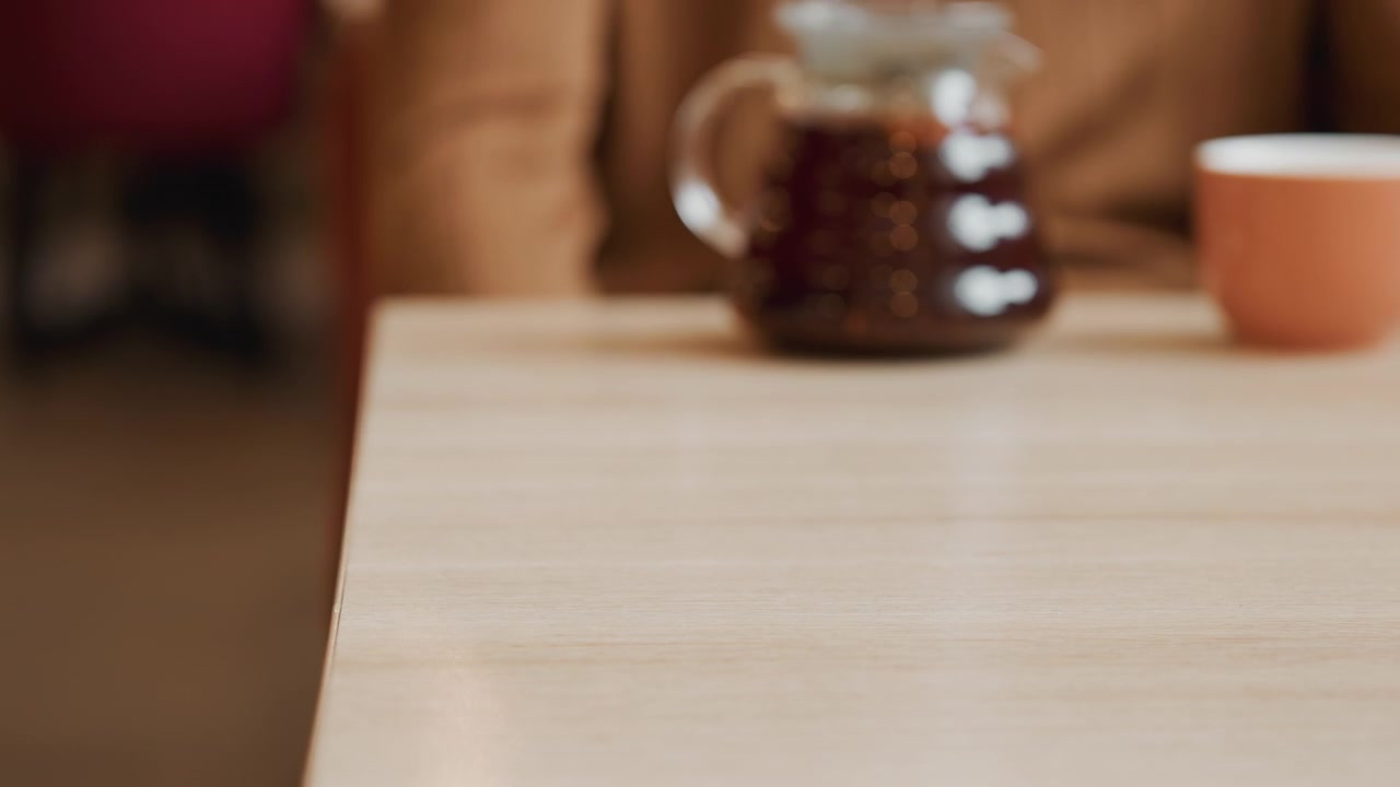 Close-up de mão movendo e colocando suavemente uma cesta de vime vazia sobre uma mesa de madeira clara. O fundo inclui uma cafeteira e xícara desfocadas, em um ambiente interno acolhedor com iluminação suave.