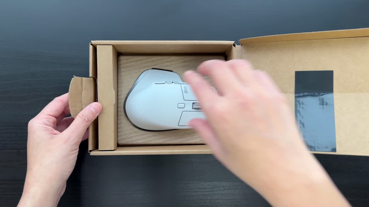 Hands open cardboard box, reveal and remove wireless ergonomic mouse on black desk, overhead view