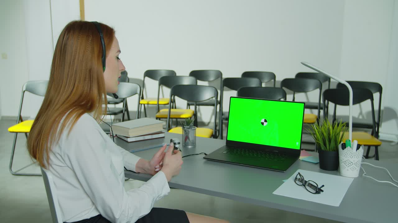 Woman in headphones talking to students via video conference at empty class. Smiling female school teacher having online class indoors. Green Screen Chroma Key Laptop for Learning. Education concept. Quarantine concept. Study by internet.