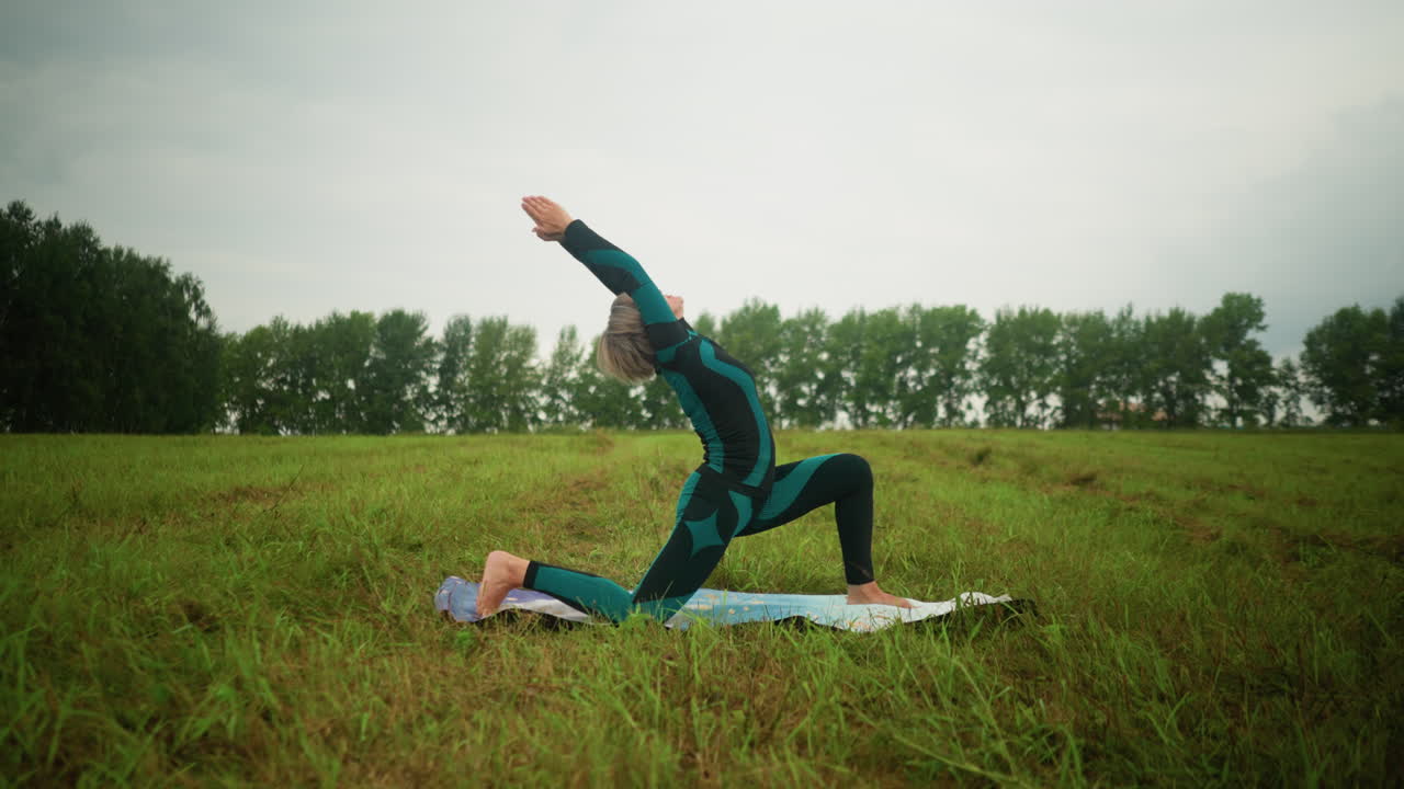 mujer de mediana edad con cabello gris practicando anjaneyasana postura de yoga baja en una alfombra de yoga en un vasto campo de hierba, con árboles alineados en la distancia bajo un cielo nublado
