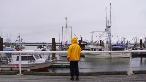 medium shot of a person in a yellow jacket walks dockside overlooking a pier of fishing boats