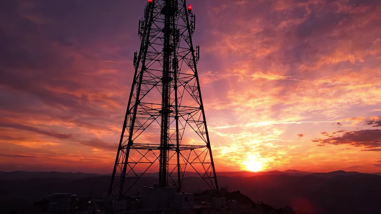 Tramonto drammatico che si affaccia su cime montuose con la silhouette di una torre di comunicazione contro i colori vibranti del cielo.