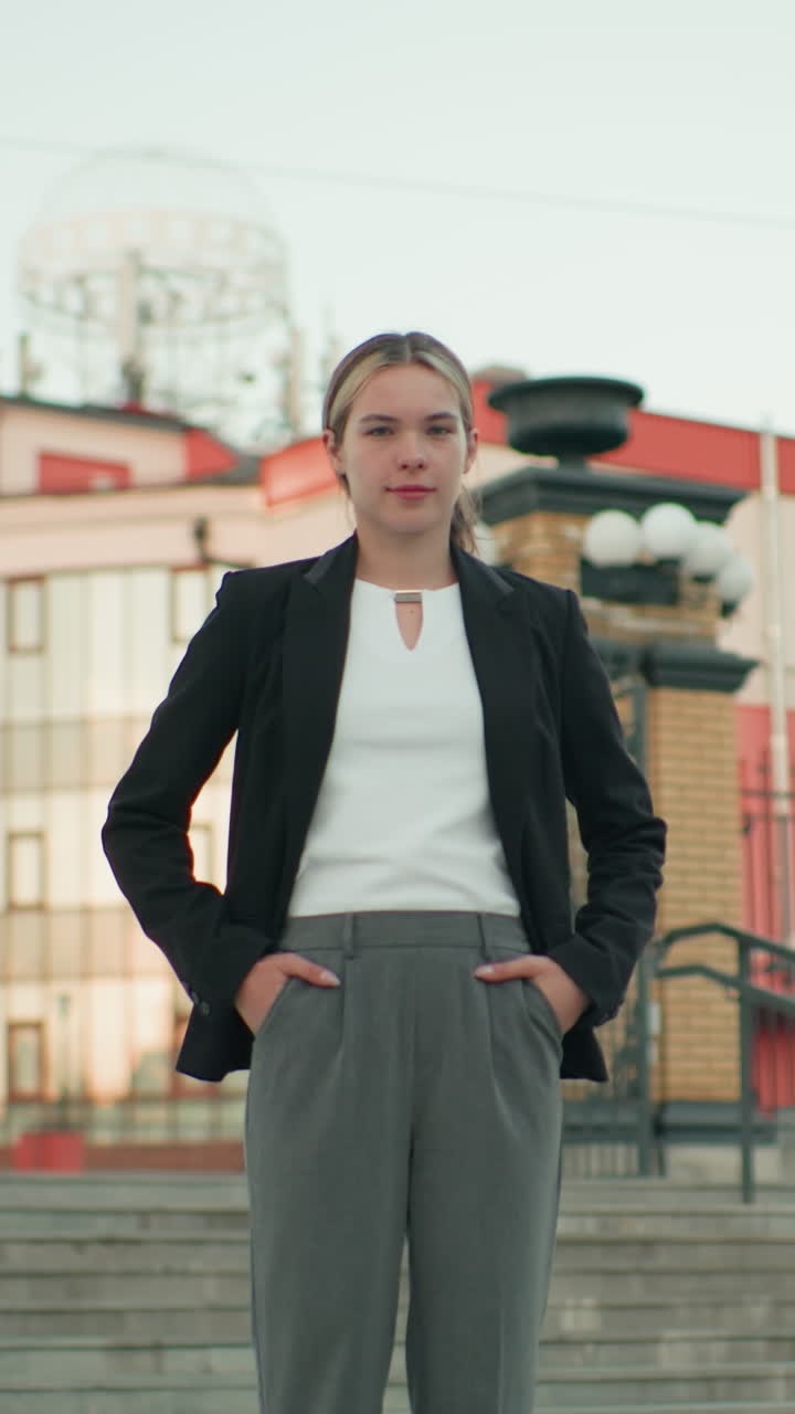 Young lady in professional attire stands confidently with hands on waist in urban setting, posing on steps in front of ornate building with metal railings and brick pillars during bright clear day