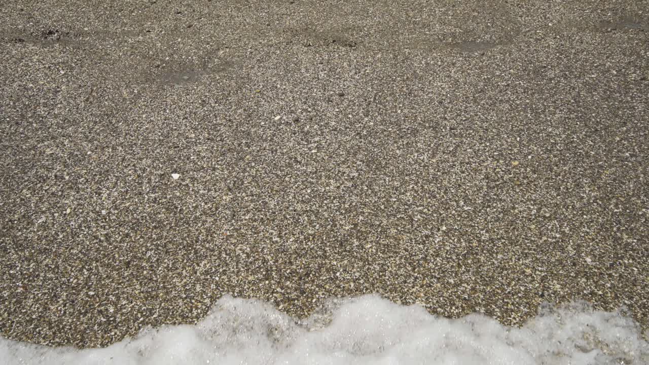 Gentle waves wash in and out over grainy sand on the sunny beach as a mans feet walking past, sinking slightly in the wet sand.