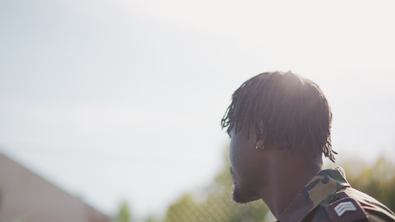 Calm Soldier Gazes Beyond Fence, Serene Armed Figure Stands Alert Amidst Suburban Scenery, Tranquil Military Personnel Examines Distant Landscape With Gentle Sunlight And Camouflage Attire