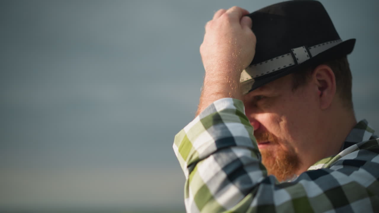 A close-up shot of a man's face, wearing a black hat with a contemplative expression, set against a backdrop of a cloudy sky. The scene captures a thoughtful moment