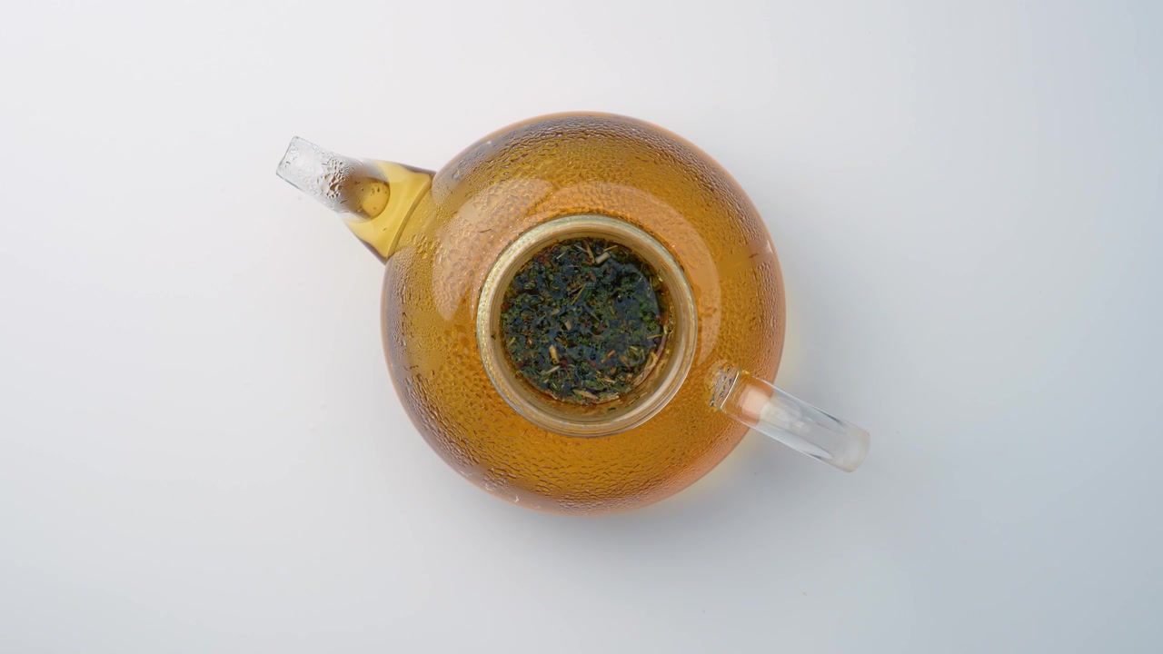 Acqua calda in infusione per tisana alla melissa in teiera. Vista dall'alto, teiera di vetro aperta con erbe verdi che galleggiano in liquido caldo su sfondo bianco liscio