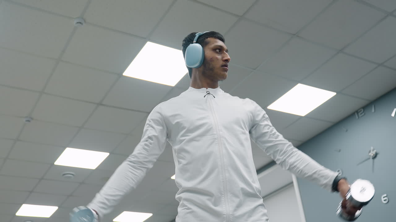 determined athlete in white jacket lifting chrome dumbbells during arm training session under bright ceiling lights, wearing wireless headphones, focusing on building upper body