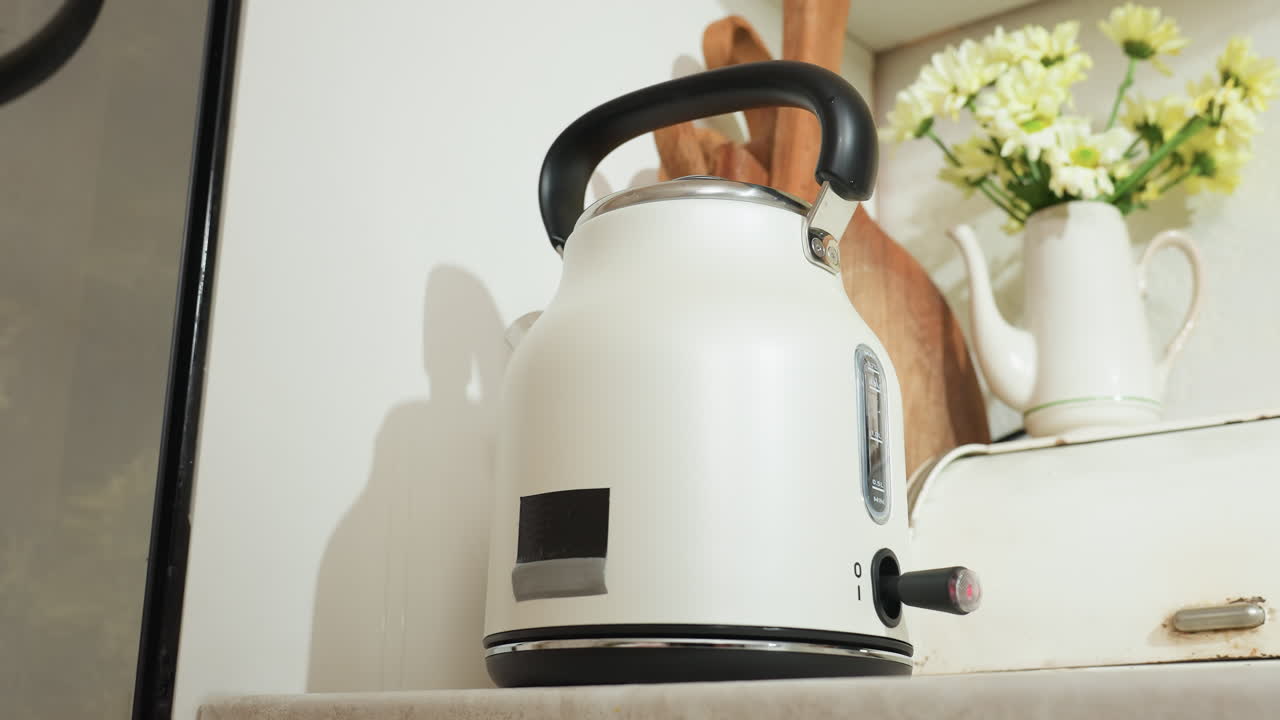 White electric kettle with water steaming inside sits on kitchen counter beside wooden spatula and ceramic flower jug as red indicator blinks and appliance gently shakes during boiling process