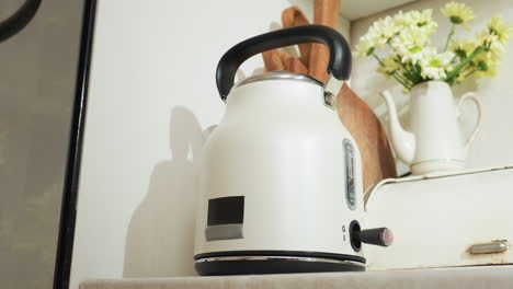 white electric kettle with water steaming inside sits on kitchen counter beside wooden spatula and ceramic flower jug as red indicator blinks and appliance gently shakes during boiling process