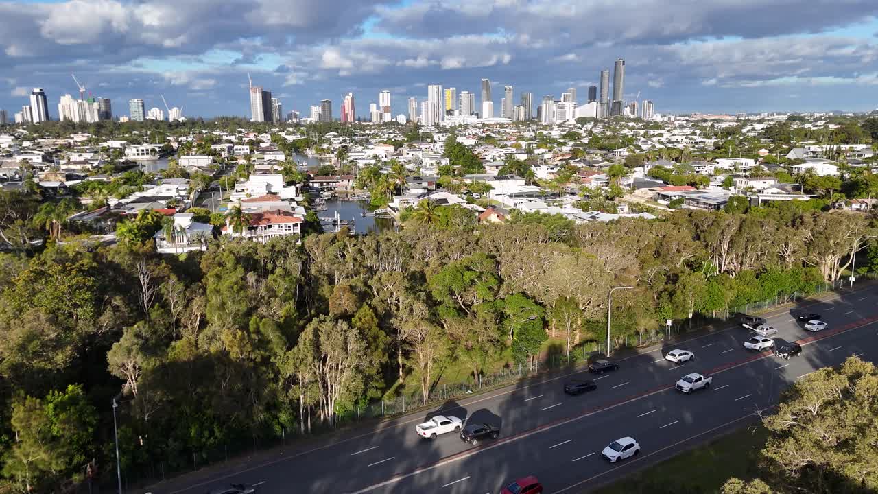 Aerial view of Gold Coast's highway, lush greenery, and canal houses under bright daylight with city skyline