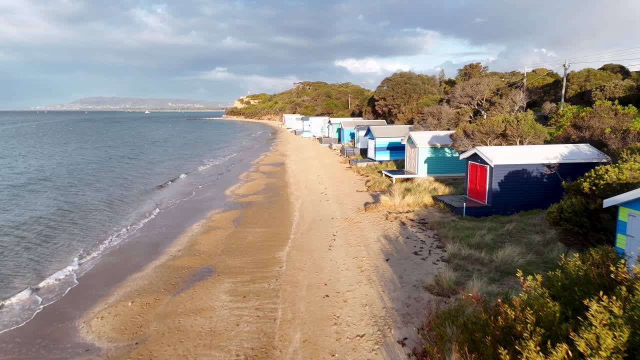 Drone glides above sandy beach, vibrant bathing boxes, and coastal vegetation under soft daylight
