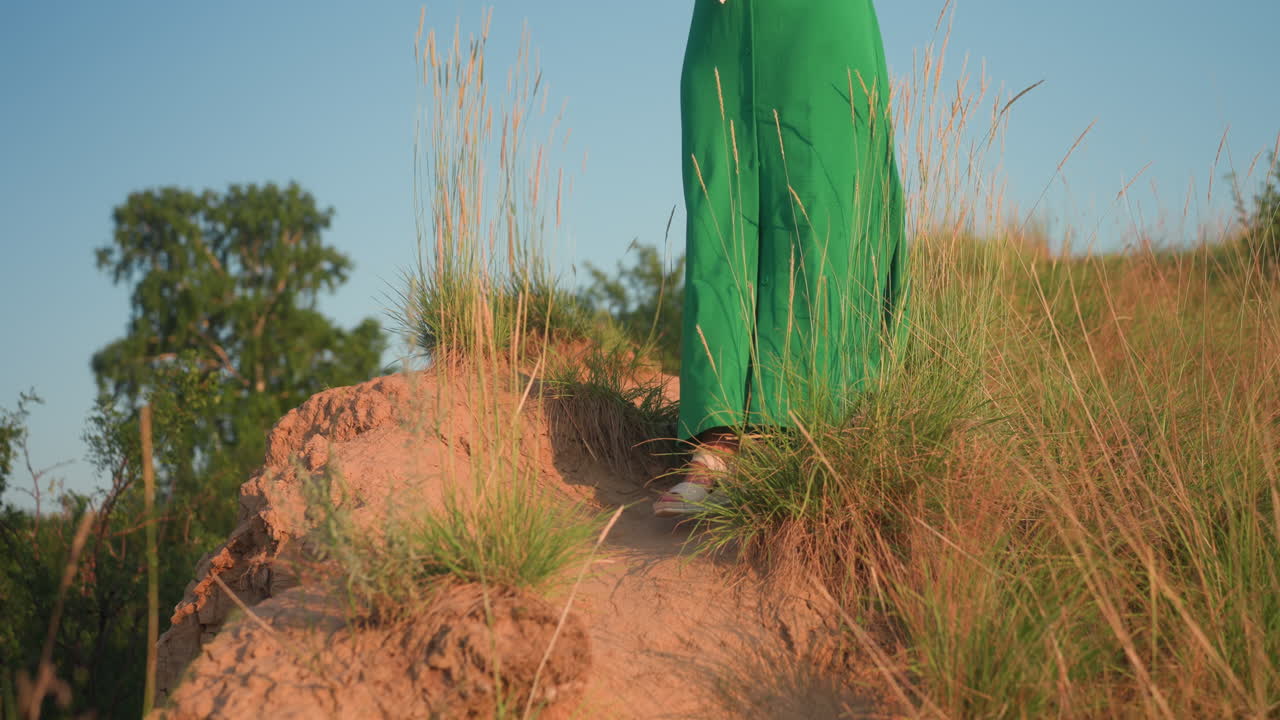 Woman walking down grassy hillside adjusting hair under warm sunset light, green dress flowing in gentle breeze as she descends slope with bubble container in hand