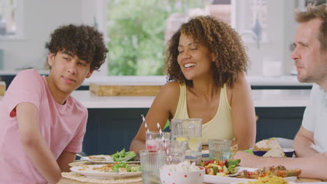 multi-racial family sitting around table in kitchen at home eating meal together