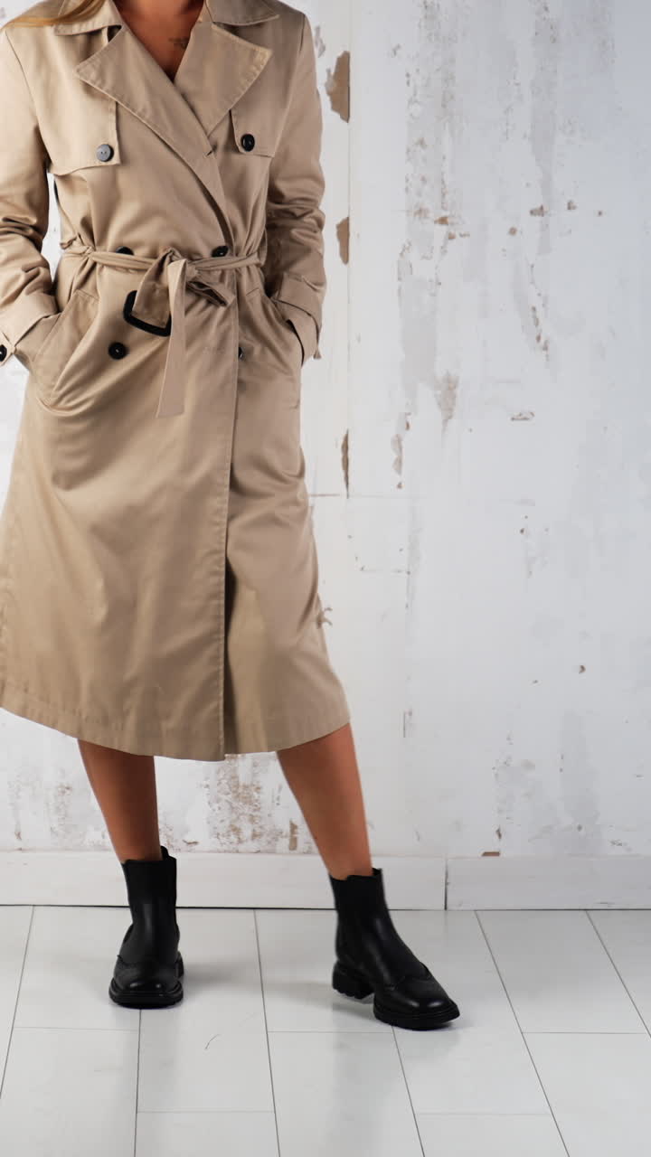 Female wearing beige coat and black boots walks in studio. Fashion and style concept. White wall backdrop. Vertical video
