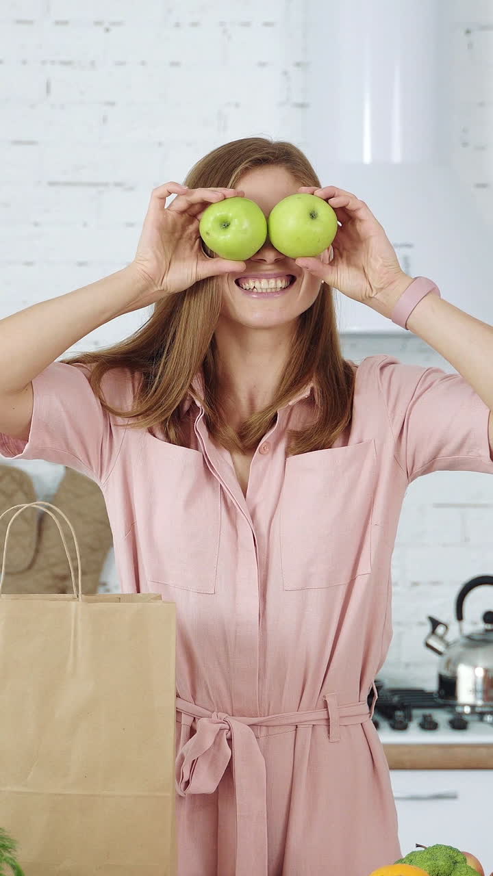 A smiling girl in the kitchen takes apples from a paper bag and puts it on her eyes. Vertical video