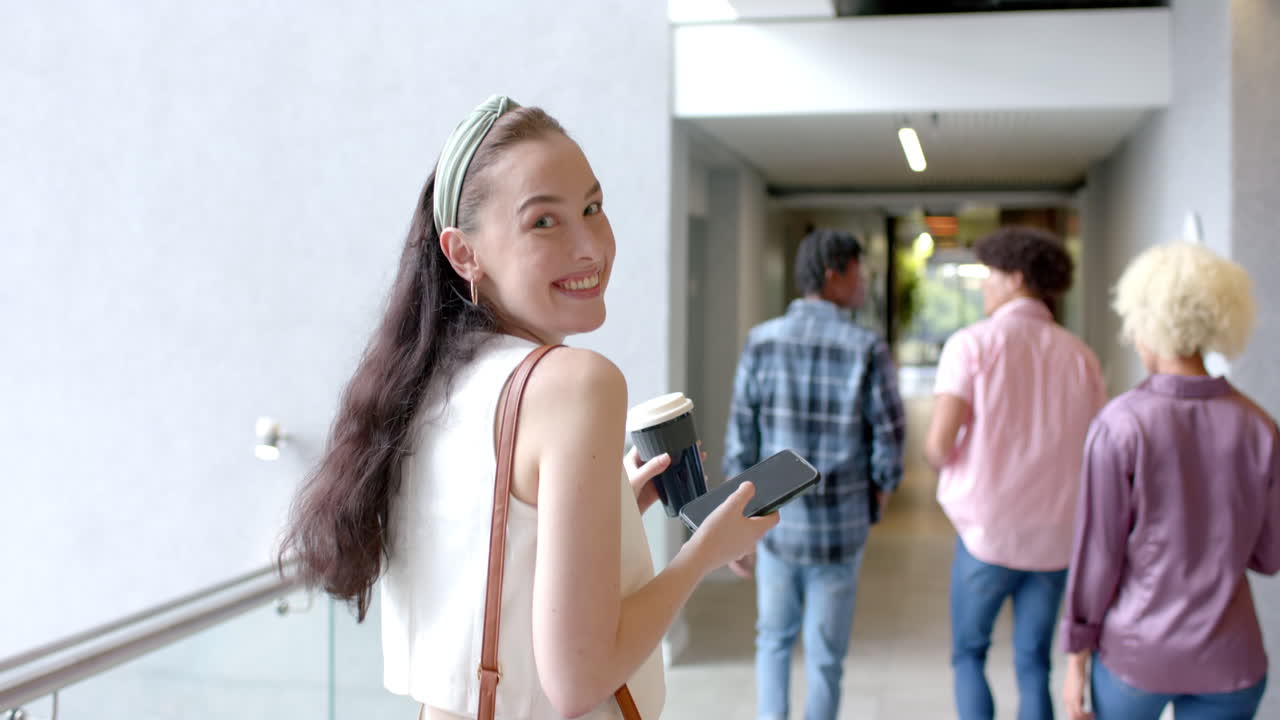 Walking in hallway, woman holding coffee cup and smartphone, smiling at camera