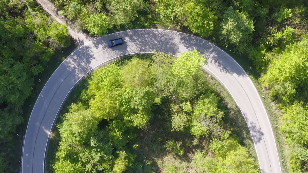 Droneshot of a semicircle curve while two cars are driving though it in a green forest, top shot aerial.