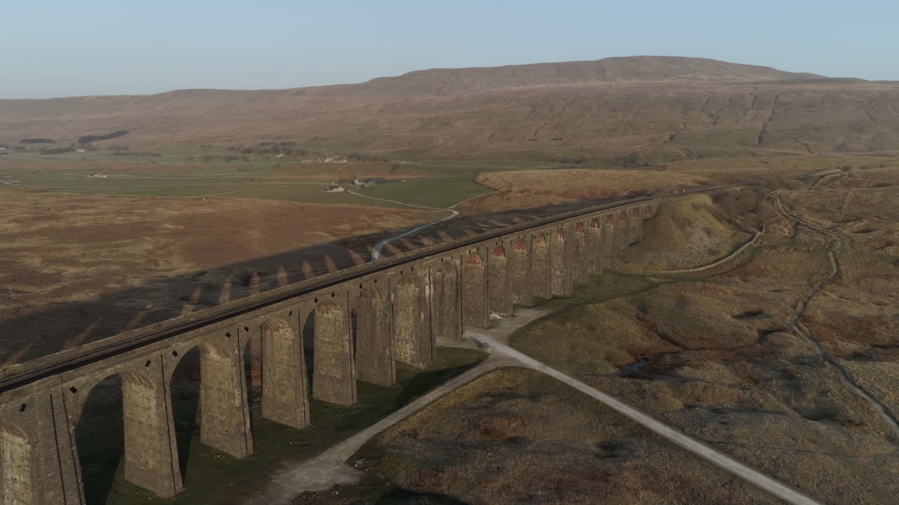 Ripresa aerea con drone in allontanamento dal ponte ferroviario del Viadotto di Ribblehead a un'alba mozzafiato, con lunghe ombre in estate, nello Yorkshire Dales, Inghilterra, Regno Unito, con la montagna Whernside (3 Cime) sullo sfondo.