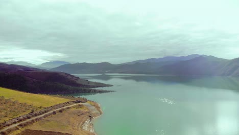 Fog-over-mountain-lake-washing-shiny-slope-hills-by-sun-rays-of-morning-on-a-peaceful-view-landscape,-aerial-shot