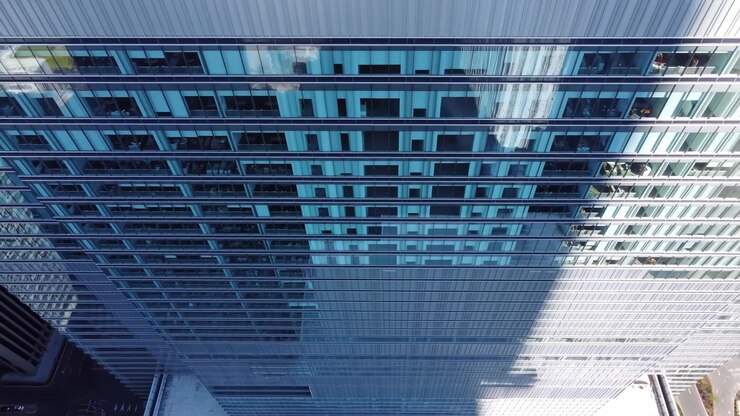 Vista aérea de la fachada de un rascacielos de cristal con ventanas reflectantes, sombra diagonal dramática, equipo en la azotea, calle