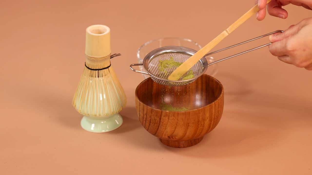 Una mano setaccia la polvere di matcha attraverso un colino in una ciotola di legno, preparazione minimalista del tè giapponese