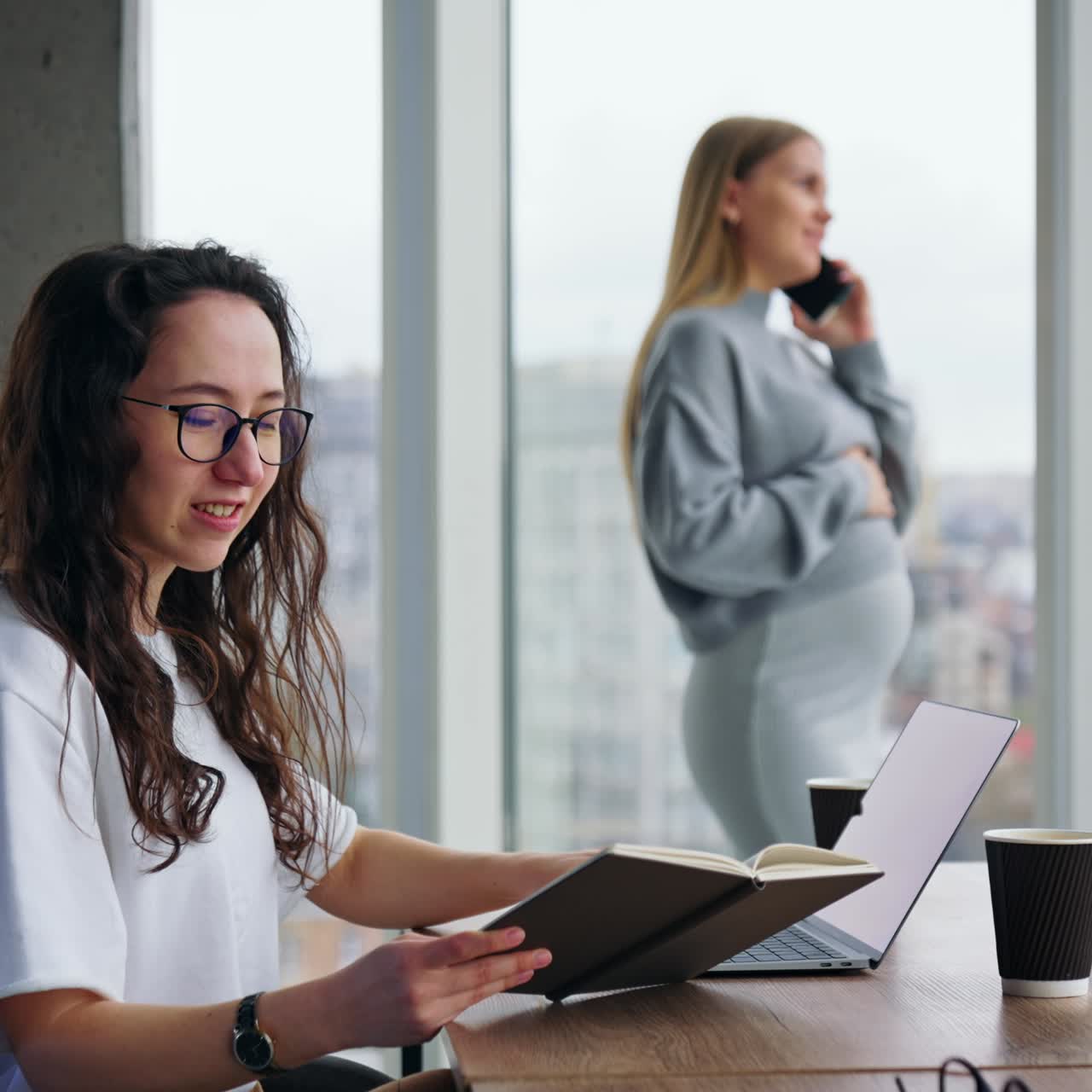 Smiling brunette looks at her notebook sitting at desk. Pregnant lady at backdrop speaks on the phone