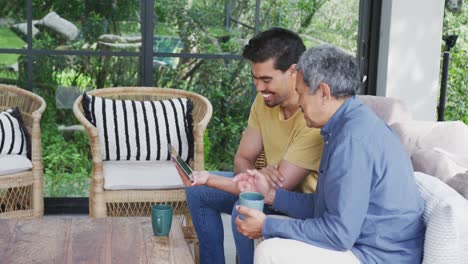 Biracial-Vater-Und-Sohn-Trinken-Kaffee,-Während-Sie-Sich-Auf-Der-Terrasse-Ein-Video-Auf-Einem-Digitalen-Tablet-Ansehen