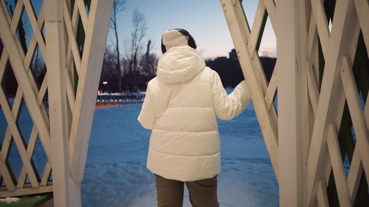 Back view of student wearing winter coat and beanie, gently swaying at gazebo entrance in snowy park during dusk twilight, hands touching lattice frame while warm ambient lights glow around