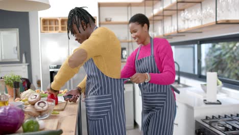 Glückliches-Afroamerikanisches-Paar-In-Schürzen,-Das-Essen-In-Der-Küche-Zubereitet,-Zeitlupe