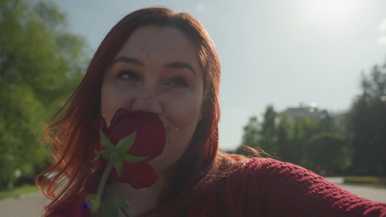 Close up of woman holding red rose and taking selfie outdoors with smartphone. Woman wears maroon knit sweater sunlight creates lens flare in background featuring green trees and clear sky