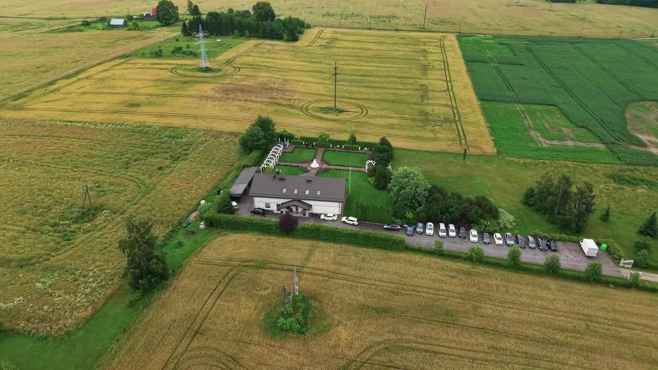 Un video con drone riprende una tenuta rurale circondata da campi agricoli dorati e verdi, con diverse auto parcheggiate, motivi geometrici di coltivazioni e alberi rigogliosi sotto una luce diurna soffusa in un'ampia ripresa aerea