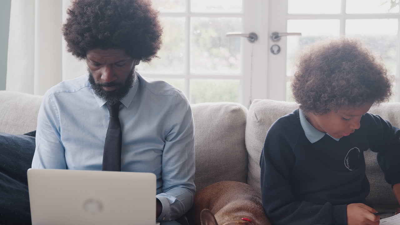 Middle aged black father and his mixed race pre teen son sitting on the sofa at home working, their pet dog sitting between them, panning shot