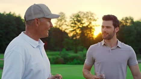 smiling golf group talking on sunset fairway. two players enjoy drink on course.