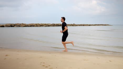 At-the-beach-along-the-edge-of-the-water-a-male-jogger-runs,-slow-motion