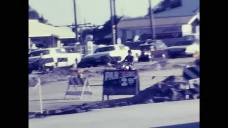 man riding motorcycle down street in 1970s usa. circa usa - 1970s: a man cruises down a street on a motorcycle in 1970s usa.