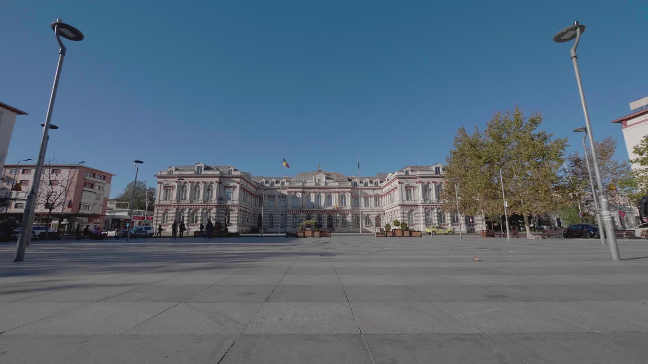 A cinematic, slow-motion dolly shot moves across a public square towards the historic Administrative Palace in Bacau, Romania, a grand neoclassical government building