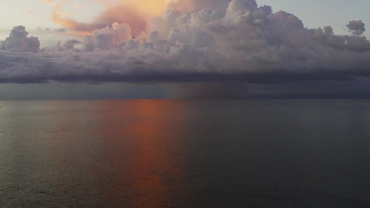 Aerial drone view of sunset in Bali with dramatic clouds over calm ocean water. Warm light reflecting on sea surface creating peaceful mood and scenic travel atmosphere