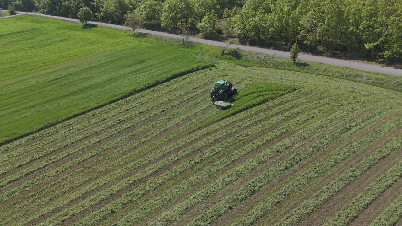 Tractor mowing green field in curved rows near roadside and tree line, drone shot.