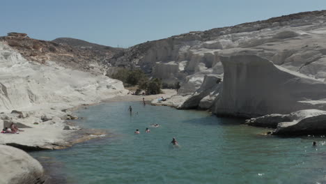 Aerial-flight-Through-Beautiful-Sarakiniko-Lunar-Volcanic-Beach-Canyon-with-Tourists-in-Water-in-Milos-Island,-Greece