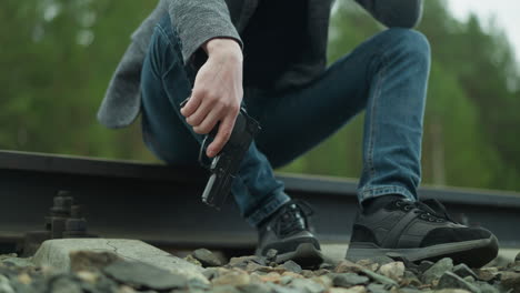 una vista cercana de una persona con una chaqueta gris, pantalones vaqueros azules y zapatos de lona negra, sentada en una vía ferroviaria, el individuo sostiene una pistola, con una vista borrosa de un bosque verde