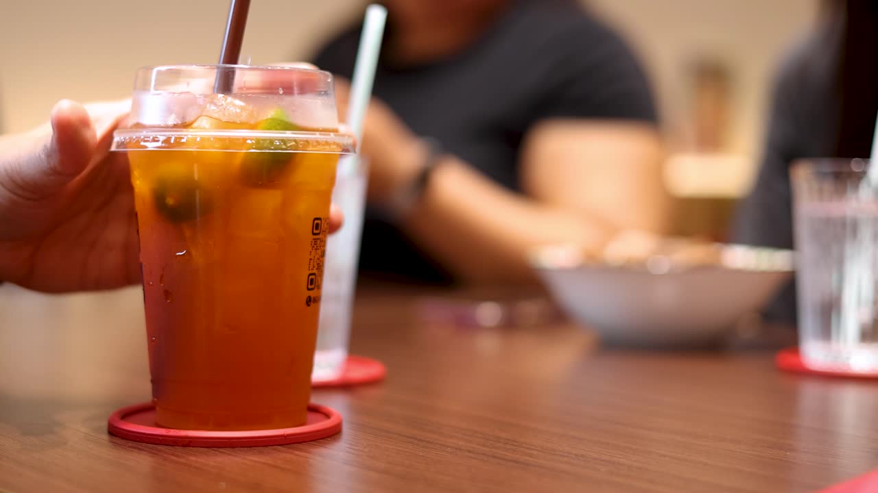 A hand lifts a plastic cup of iced lemon tea from a table in a warmly lit, casual dining environment, with blurred background figures and tableware