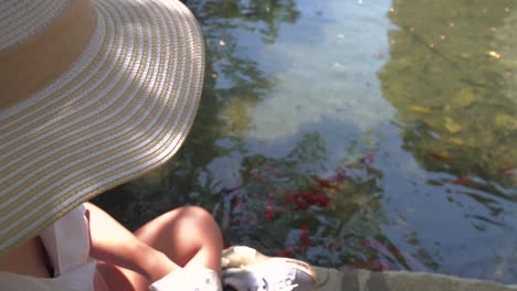 top down view of girl in summer hat giving food to koi fish in pond with reflections